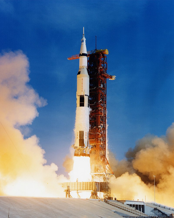 A photograph of the Apollo 11 Saturn V rocket at the moment of launch. The rocket is producing an enormous bright plume and has risen slightly off the launch platform.