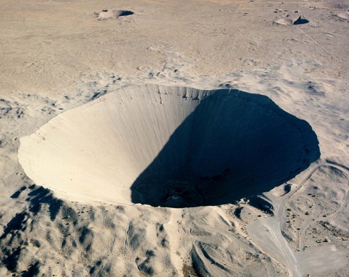 An image of the crater left by the Sedan test. The crater is a large conical hole of more or less uniform beige dirt.