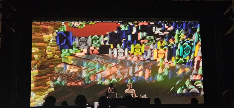 Photo of Lucid and Kahtrin Hunze sitting with laptops and mixers in front of a gigantic splash of colourful shapes projected over and behind them.