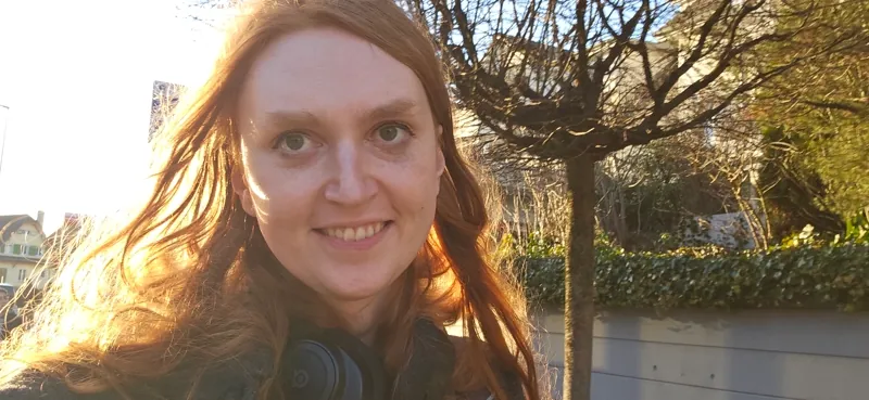 A picture of me, a trans woman with red hair, smiling while backlit by the sun, blowing out the camera a bit.