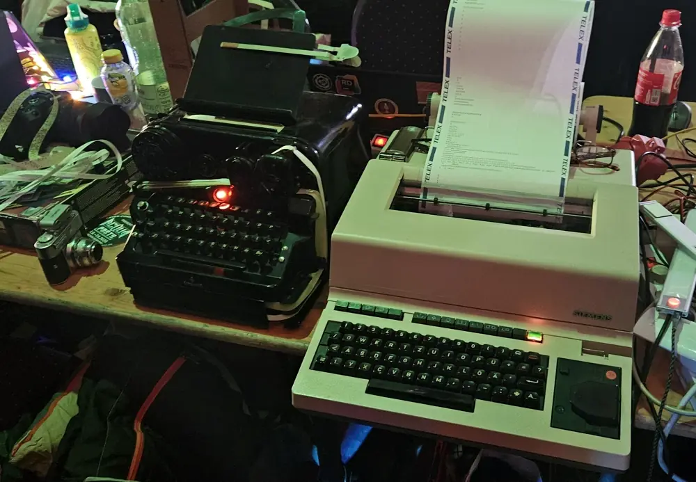 An old black, metal German Teletype machine next to a white plastic Sieman Telex machine.